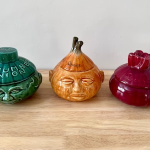 May include: Three ceramic lidded jars shaped like vegetables with human faces. The green jar is labelled CUCUMBER ONION, the orange jar is labelled ONION, and the deep red jar is labelled BEETROOT. Each jar features a sculpted face and lid.