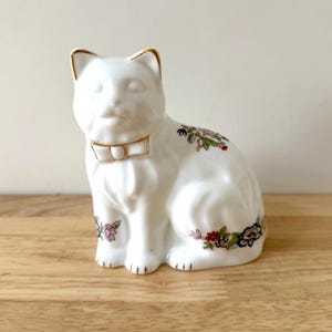 May include: A white ceramic cat figurine with gold trim on the ears and bow tie. The cat is decorated with floral patterns in pink, green, and blue. The figurine is sitting on a wooden surface.