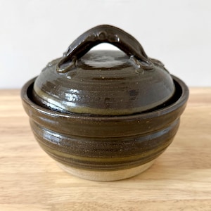 May include: A handmade ceramic lidded bowl in shades of brown and tan. The bowl has a rounded shape with a lid and a handle. The exterior features horizontal grooves and a glossy finish, perfect for kitchen decor.