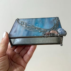 May include: A rectangular stained glass box with a blue and white marbled design on the lid. The box is trimmed with silver metal and features decorative stones and a silver metal design on the top. The box is held in a hand.