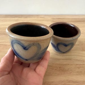 Op de afbeelding: Twee kleine, bruine steengoed potten met blauwe hartmotieven. De potten zijn geglazuurd en hebben een rustieke uitstraling.