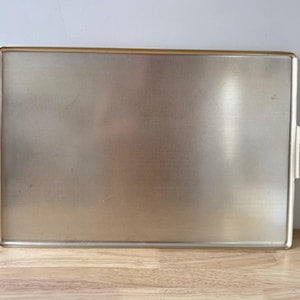 May include: A rectangular metallic serving tray with a gold-toned finish and a fine, textured crosshatch pattern across the surface. The tray features two integrated side handles with a matching gold finish and a raised rim around the edge.