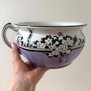 May include: A white ceramic chamber pot with a black and white floral design and a lavender glaze. The pot has a single handle.