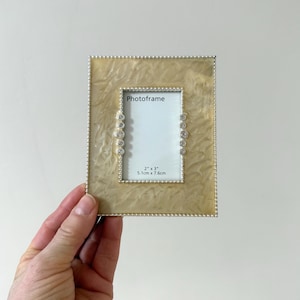 May include: A square photo frame with a pearlescent finish and a beaded border. The frame has a rectangular opening for a 2" x 3" (5.1cm x 7.6cm) photo. The frame is embellished with decorative rhinestones.