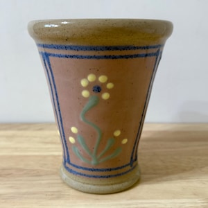 May include: A handcrafted ceramic vase with a flared design. The vase has a brown exterior with a pink central panel. A blue border frames the panel, and a floral design with yellow and green accents decorates the front. The rim and base are a lighter brown.