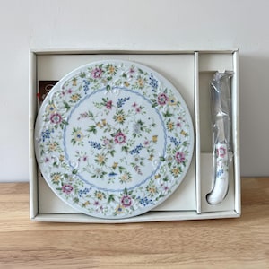 May include: A white ceramic cheese board and knife set. The board features a floral pattern with pink, yellow, and blue flowers. The knife has a matching floral design on the handle. The set is presented in a white box.