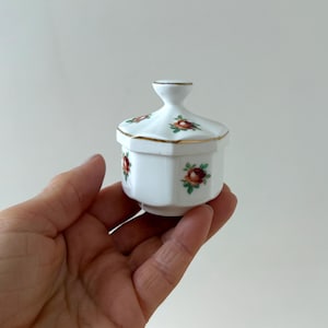 May include: A small white porcelain trinket box with a lid. The box is decorated with a gold trim and red and green floral designs.