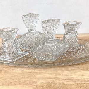 Op de afbeelding: Een doorzichtige glazen toilettafelset met een ruitpatroon. De set bevat een rechthoekige dienblad, twee kleine kandelaars en twee parfumflesjes. De items worden gepresenteerd op een houten oppervlak.