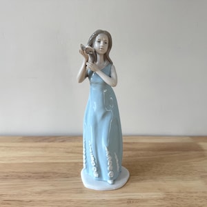 May include: A porcelain figurine of a woman in a light blue dress, holding a seashell to her ear. The figurine has a white base and is approximately 10 inches tall. The dress has decorative white accents.