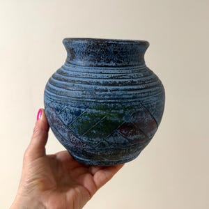 May include: A blue ceramic vase with a textured surface and a woven pattern on the lower half. The vase has a round body and a narrow neck. The color is a mix of blue, green, and red.