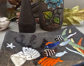 May include: A collection of handcrafted ceramic pieces. Includes a dark brown vase, a tall bottle with a blue floral design, and a square dish with colorful fish and starfish motifs. The pieces have a rustic, textured finish.