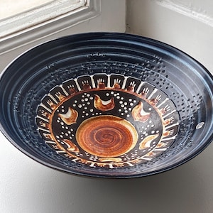 May include: A large, round ceramic bowl with a dark blue and black exterior. The interior features a central brown spiral, surrounded by a ring of stylized white and brown bird motifs, white dots, and a decorative border of vertical white lines.