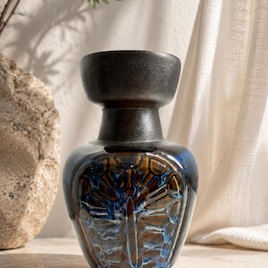 Peut inclure: Un vase en céramique avec un bord et un col brun foncé, passant à un corps brillant et orné de motifs bleus, bruns et blancs. Le vase présente un design texturé unique et repose sur une surface claire.