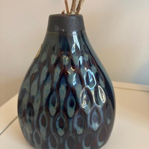 Puede incluir: Un jarrón de cerámica azul oscuro con una superficie texturizada y estampada. El jarrón tiene un cuerpo redondo y un cuello estrecho. Se insertan tallos de plantas secas en el jarrón. El jarrón está sobre una superficie blanca.