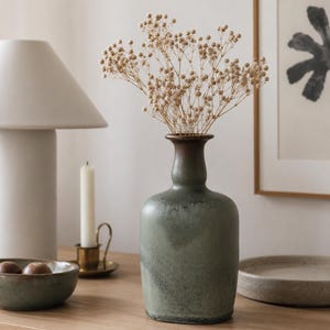 May include: A ceramic vase with a mottled sage green and dark brown glaze, featuring a rounded body and a narrow neck. The vase contains a cluster of dried, light beige baby's breath flowers, which extend upwards from the opening of the vessel.