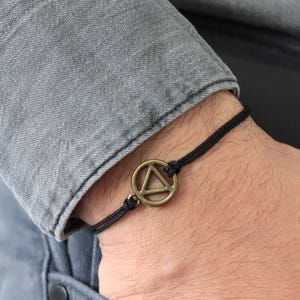 Può includere: Un braccialetto con cordino nero e ciondolo a triangolo color bronzo. Il braccialetto è indossato su un polso, con una camicia di jeans grigia e jeans sullo sfondo. Il ciondolo è un semplice design geometrico.