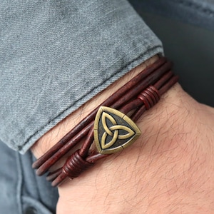 May include: A brown leather bracelet with a bronze-colored Celtic knot charm. The bracelet features multiple strands of dark brown leather and is secured with a clasp. The bracelet is worn on a wrist.