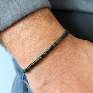 May include: A beaded bracelet featuring alternating dark green and black beads. The bracelet is worn on a wrist, with the arm in a gray shirt and blue jeans. The bracelet is a simple, minimalist accessory.