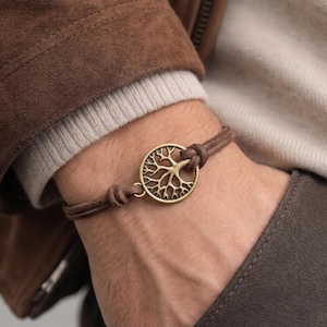 Puede incluir: Una pulsera de cuero marrón con un dije de árbol de la vida de bronce. La pulsera se lleva en la muñeca, con una chaqueta marrón y un suéter beige en el fondo. El dije es un diseño circular detallado.