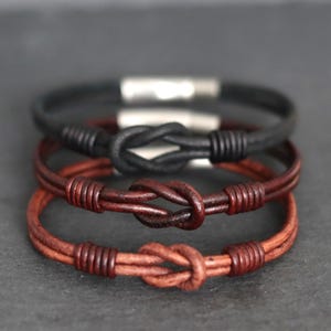 May include: Three leather bracelets in black, dark brown, and reddish-brown. Each bracelet features a knot design in the center and a silver clasp. The bracelets are stacked on top of each other.