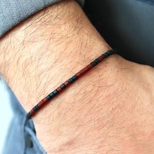 May include: A beaded bracelet with a minimalist design. The bracelet features small, cylindrical beads in alternating colors of black and reddish-brown. The bracelet is worn on a wrist, with a gray shirt and blue jeans in the background.