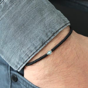Pode incluir: Pulseira de contas pretas com uma única conta azul clara. A pulseira é usada em um pulso, com uma camisa jeans cinza e jeans azuis ao fundo. A pulseira tem um design simples e minimalista.