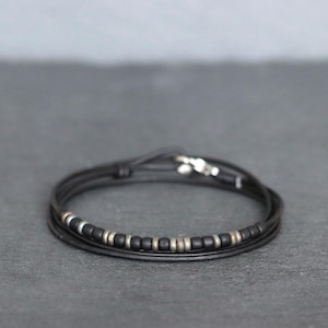 May include: Black leather bracelet with a wrap-around design. The bracelet features small, black beads and silver-coloured beads. The clasp is silver. This minimalist bracelet is suitable for everyday wear.