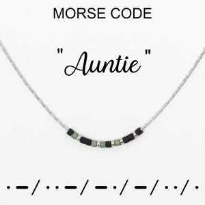 Könnte beinhalten: Eine silberne Halskette mit einem Anhänger, der "Auntie" in Morsecode buchstabiert. Der Anhänger hat kleine schwarze und grüne Perlen. Die Wörter "MORSE CODE" und die Morsecode-Übersetzung befinden sich oberhalb und unterhalb der Halskette.