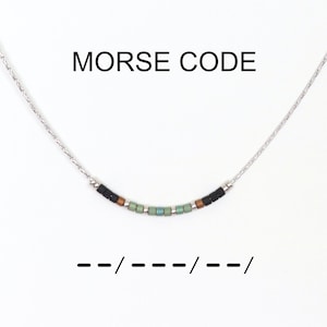 Op de afbeelding: Zilveren ketting met een gebogen staafhanger. De hanger heeft een Morsecode-ontwerp met zwarte, bruine en groene kralen. De tekst "MORSE CODE" staat boven de ketting.