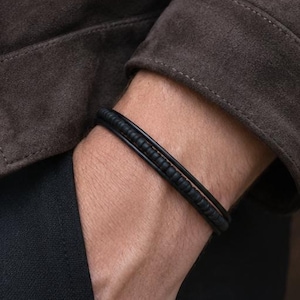 May include: A black leather bracelet with a braided design is worn on a wrist. The bracelet is the focal point, set against a dark brown suede jacket and black pants. The image is a close-up, highlighting the bracelet's texture and style.