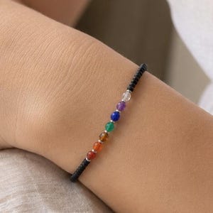 May include: A black beaded bracelet with seven colorful gemstone beads. The bracelet is worn on a wrist, with a white top in the background. The gemstones are in the colors of the chakras.