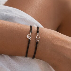 Könnte beinhalten: Zwei schwarze Kordelarmbänder mit silberfarbenen Anhängern am Handgelenk. Einer ist herzförmig, der andere eine Flaschenform. Die Person trägt ein weißes Kleid und sitzt am Meer. Schlichtes, elegantes Design.