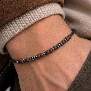 Può includere: Un braccialetto con perline rosso scuro e argentate infilate su un cordino nero. Il braccialetto è indossato su un polso, con una giacca marrone e un maglione beige sullo sfondo.