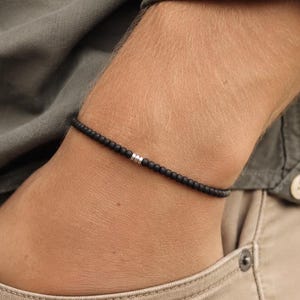 Puede incluir: Una pulsera fina compuesta por pequeñas cuentas negras uniformes dispuestas en una banda circular continua. Una única cuenta cilíndrica de metal en tono plateado se sitúa como pieza de acento central en la hilera de cuentas negras.