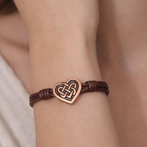 May include: A brown leather bracelet with a copper heart-shaped charm. The charm features an intricate Celtic knot design. The bracelet is worn on a wrist, showcasing the jewelry's details and craftsmanship.