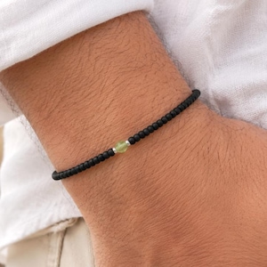 May include: Black braided bracelet with a small, light green bead. The bracelet is worn on a wrist, with a white shirt and beige trousers in the background. The bracelet is a simple, minimalist design.