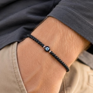 May include: A beaded bracelet featuring small, matte black spherical beads arranged in a continuous loop. A single circular charm with a blue, white, and black eye-like design is positioned at the centre, flanked by two small silver-toned metal spacers.