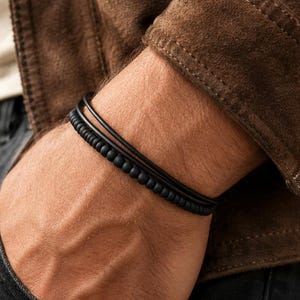 May include: A set of two black bracelets worn on the wrist. One bracelet consists of a single smooth black cord, while the other features a series of small, uniform matte black spherical beads arranged in a continuous circular pattern.