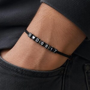 May include: Black beaded bracelet with white lettered beads spelling "S ♡ D 08 17". The bracelet is on a black cord. The person is wearing a dark grey long-sleeve shirt and dark grey jeans.