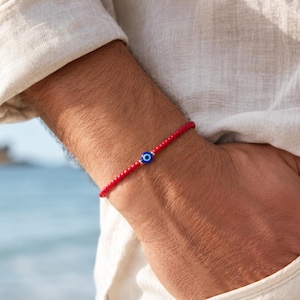 Puede incluir: Pulsera de cuentas rojas con un dije de ojo turco azul y blanco. La pulsera se lleva en la muñeca, con una camisa de color claro en el fondo. Un accesorio sencillo pero elegante.