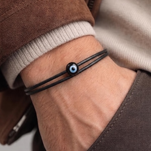 Può includere: Bracciale in pelle nera con una perla a forma di occhio blu e bianco. Il bracciale è composto da due sottili cordoncini in pelle nera e una perla rotonda blu scuro con un disegno a occhio bianco e nero. Il bracciale è indossato al polso.