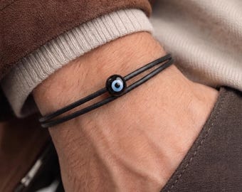 May include: Black leather bracelet featuring a blue and white evil eye bead. The bracelet is made of two thin black leather cords and a round, dark blue bead with a white and black eye design. The bracelet is worn on a wrist.