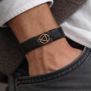 Può includere: Bracciale in pelle nera con un ciondolo geometrico color oro. Il bracciale è indossato su un polso, con una camicia bianca e un maglione grigio visibili sullo sfondo. Il bracciale è largo circa 2 cm.