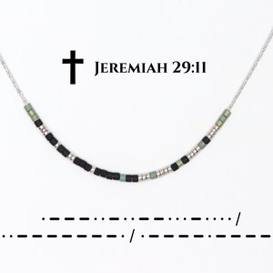 May include: A silver necklace with a beaded design. The necklace features black, silver, and green beads. The text "Jeremiah 29:11" is above the necklace, with a cross symbol. Below the necklace is a line of Morse code.