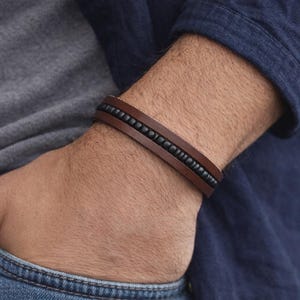 Könnte beinhalten: Braunes Lederarmband mit einer Reihe schwarzer Perlen. Das Armband wird am Handgelenk getragen, die Hand der Person in einer Tasche. Ein schlichtes, aber stilvolles Accessoire.