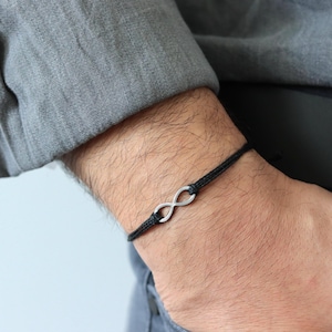 Könnte beinhalten: Ein schwarzes Kordelarmband mit einem silbernen Unendlichkeitssymbol. Das Armband wird am Handgelenk getragen, vor dem Hintergrund eines grauen Hemdes und einer blauen Jeans.