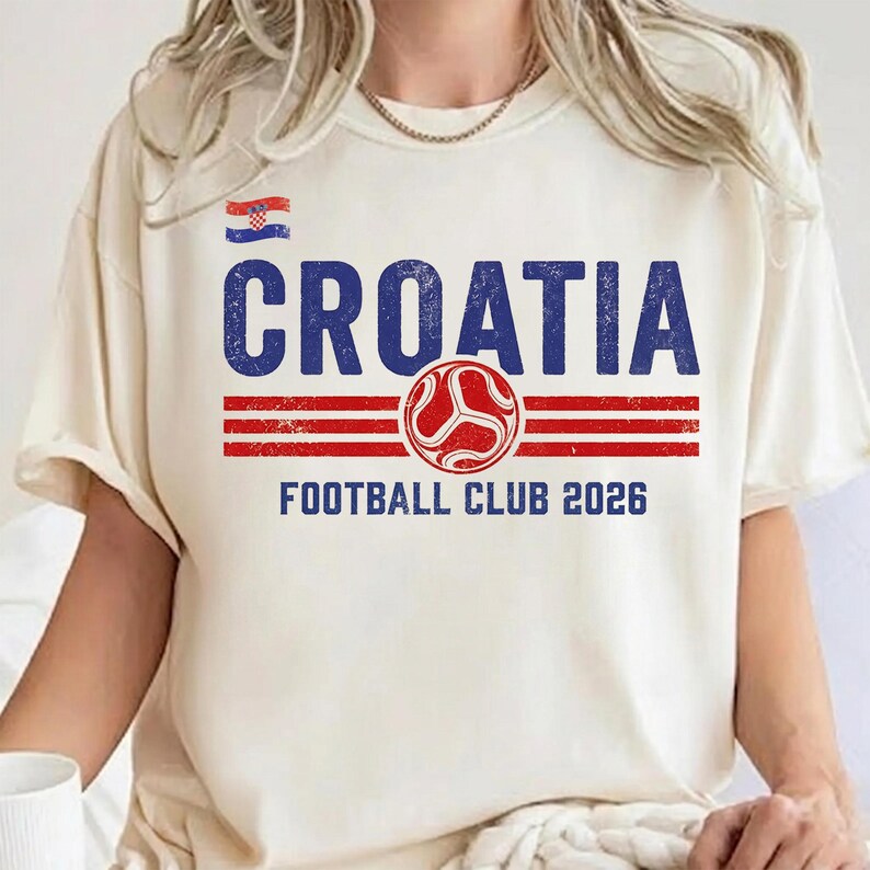 Peut inclure: Un t-shirt &agrave; manches courtes de couleur cr&egrave;me avec le texte CROATIA en bleu, un petit drapeau croate, trois bandes horizontales rouges, un embl&egrave;me circulaire rouge et le texte FOOTBALL CLUB 2026 en bleu. Le motif a un aspect vieilli et us&eacute;.