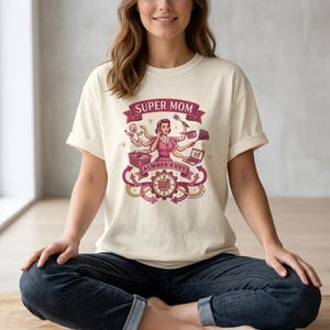 Puede incluir: Camiseta de color crema con un gráfico de una mujer con múltiples brazos, sosteniendo varios objetos como un bebé, una computadora portátil y artículos de limpieza. Los textos "SUPER MOM" y "ALWAYS BUSY" se muestran en un diseño de banner. La camiseta la lleva una persona.