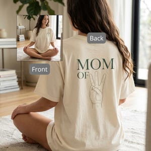 Puede incluir: Camiseta color crema con el texto "MOM OF 3" y un gesto de la mano en la espalda. La parte delantera de la camiseta tiene un pequeño diseño. La camiseta la lleva una persona sentada con las piernas cruzadas.