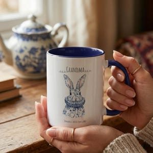 Puede incluir: Taza de cerámica blanca con interior y asa azules. La taza presenta una ilustración de un conejito con estampados florales azules y la palabra "GRANDMA" encima. Los nombres "DUNCAN & JACK & TOM & ABBY" están impresos debajo.